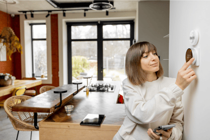 A woman in a creme-colored outfit, leaning on a wall, adjusting the settings of her wall-mounted thermostat.