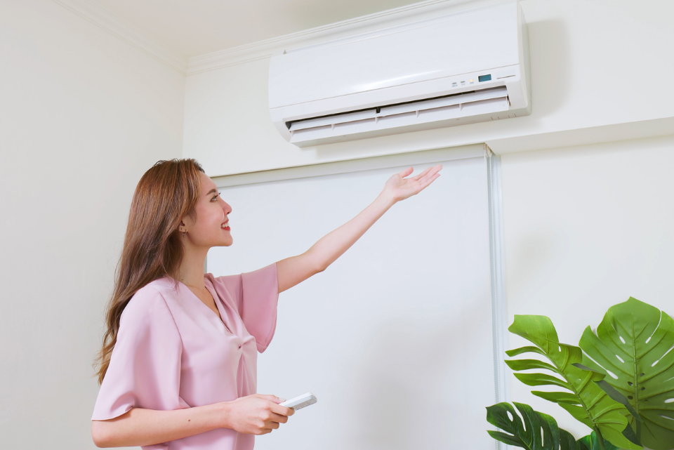 A woman stands in front of an indoor mini-split, her hand outstretched to feel the emitted fresh air.